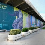 Marciapiede dell'aeroporto di Catania con fioriere e cartellonistica nelle vetrate con i principali monumenti di Catania e la scritta "Benvenuti in Sicilia"