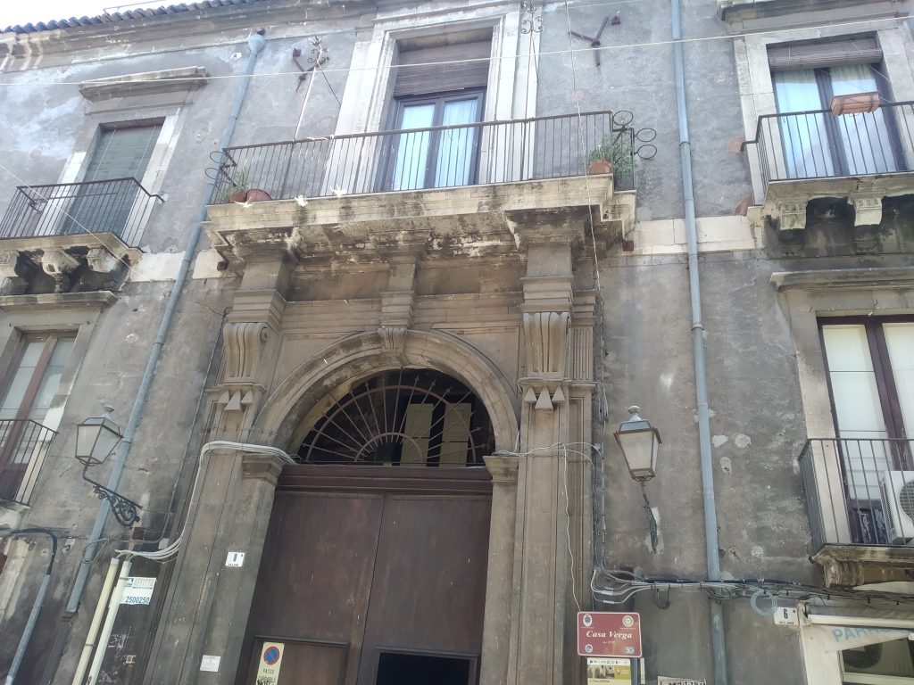 Museo Casa Verga: la facciata storica con il grande portone di legno marrone scuro e gli intonaci grigi. In alto i balconi