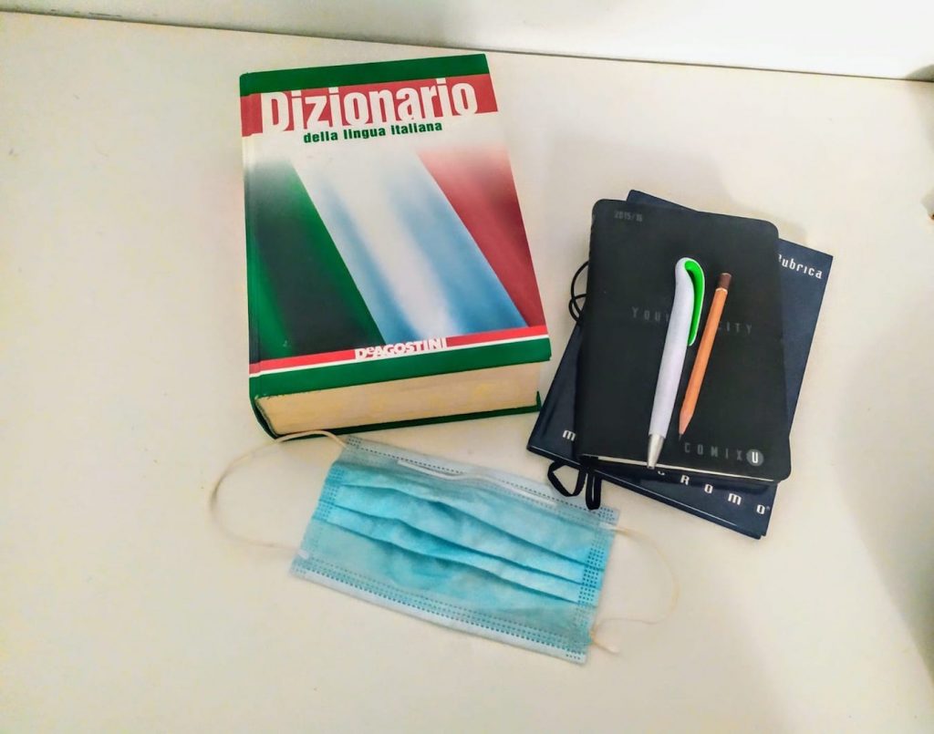 Dizionario d'italiano, rubrica, penne ,matita e mascherina blu sopra una scrivania bianca