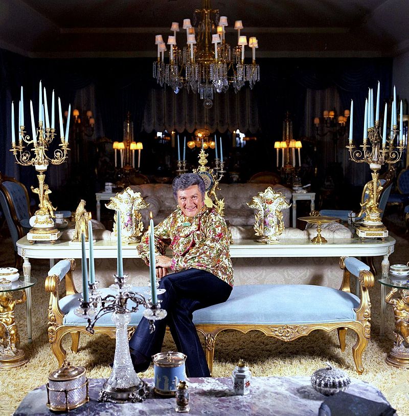 Liberace seduto su una lunga panca celeste circondato da candelabri